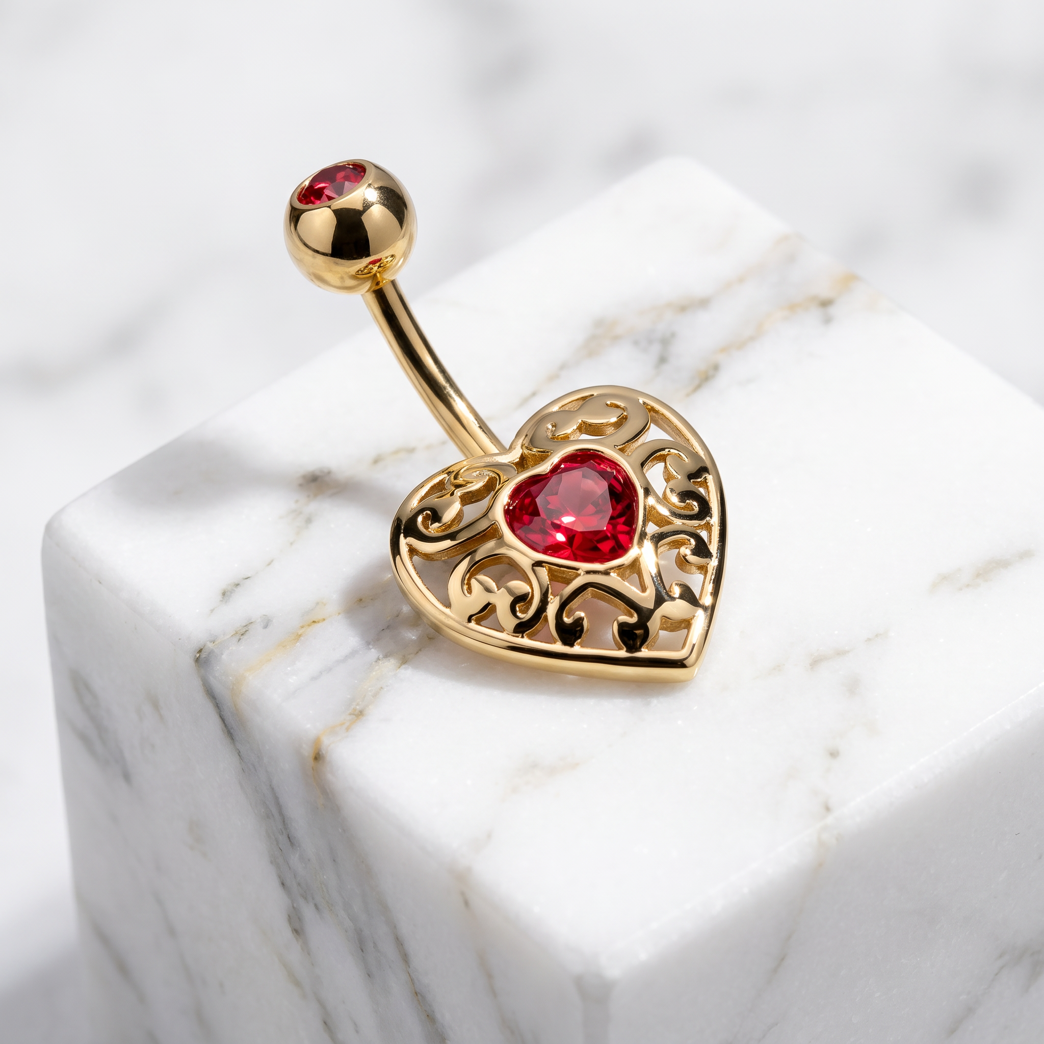 Gold-plated belly button ring with an ornate filigree heart design and a red heart-shaped gemstone, resting on a white marble cube.