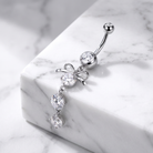 A silver-toned navel ring with a curved barbell, a round clear gemstone, a bow-shaped setting, and three dangling clear gemstones on a white marble block.