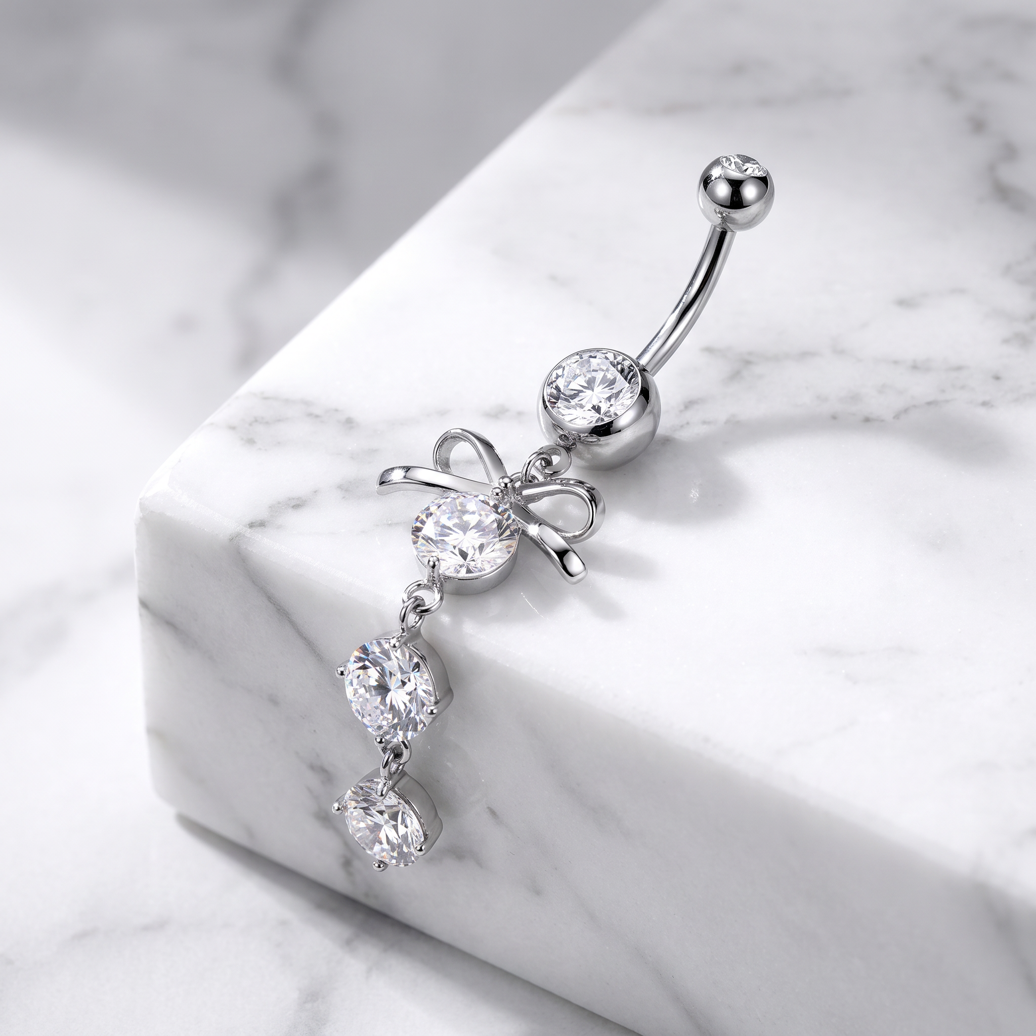 A silver-toned navel ring with a curved barbell, a round clear gemstone, a bow-shaped setting, and three dangling clear gemstones on a white marble block.