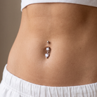 A close-up of a person's midsection showing a belly button piercing with a rose gold barbell and clear rhinestone charms, styled with a white ribbed crop top and white sweatpants.