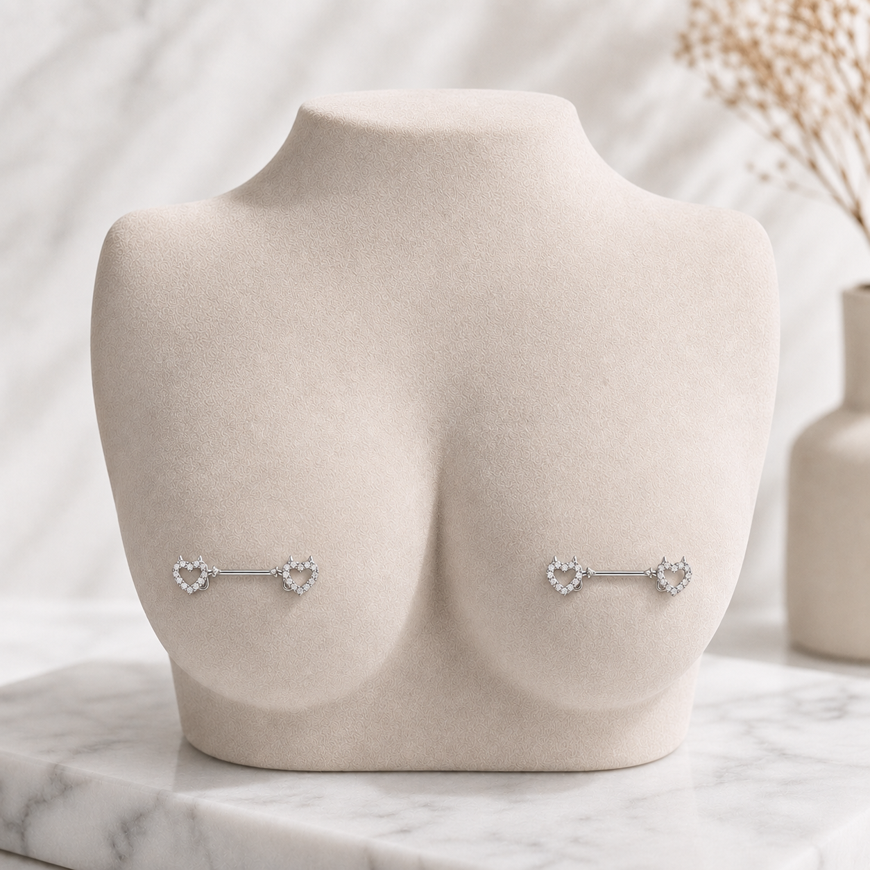 A pair of silver barbell nipple piercings with heart-shaped clear crystal clusters, displayed on a beige fabric-covered female torso mannequin against a marble and dried flower background.