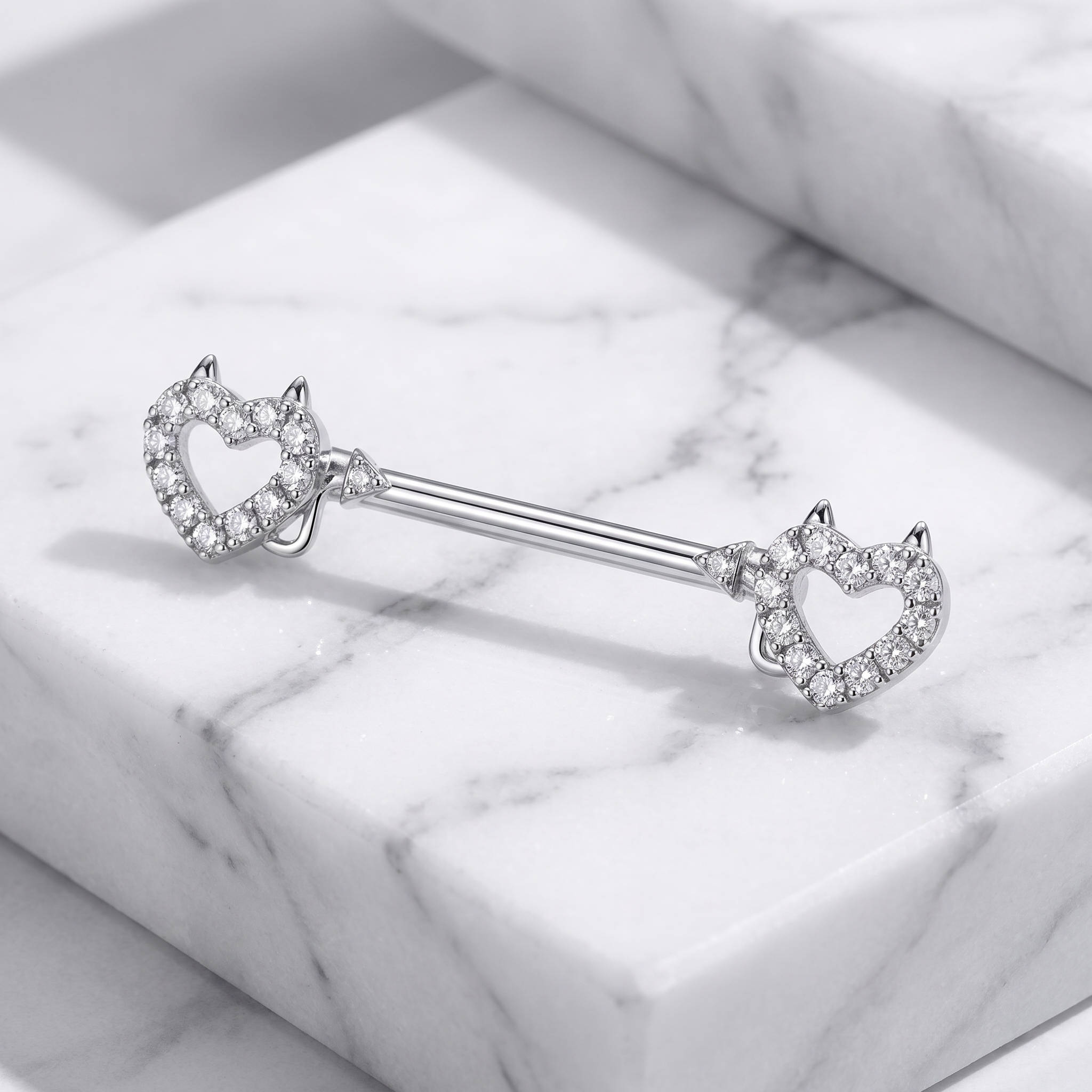 A silver-toned barbell-style body jewelry piece with two open heart-shaped ends, each outlined with small clear stones and topped with tiny pointed details, displayed on a white marble surface.