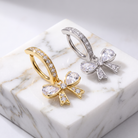 Two bow-shaped hoop earrings displayed on a white marble block. One earring is gold with clear stones, and the other is silver with clear stones.