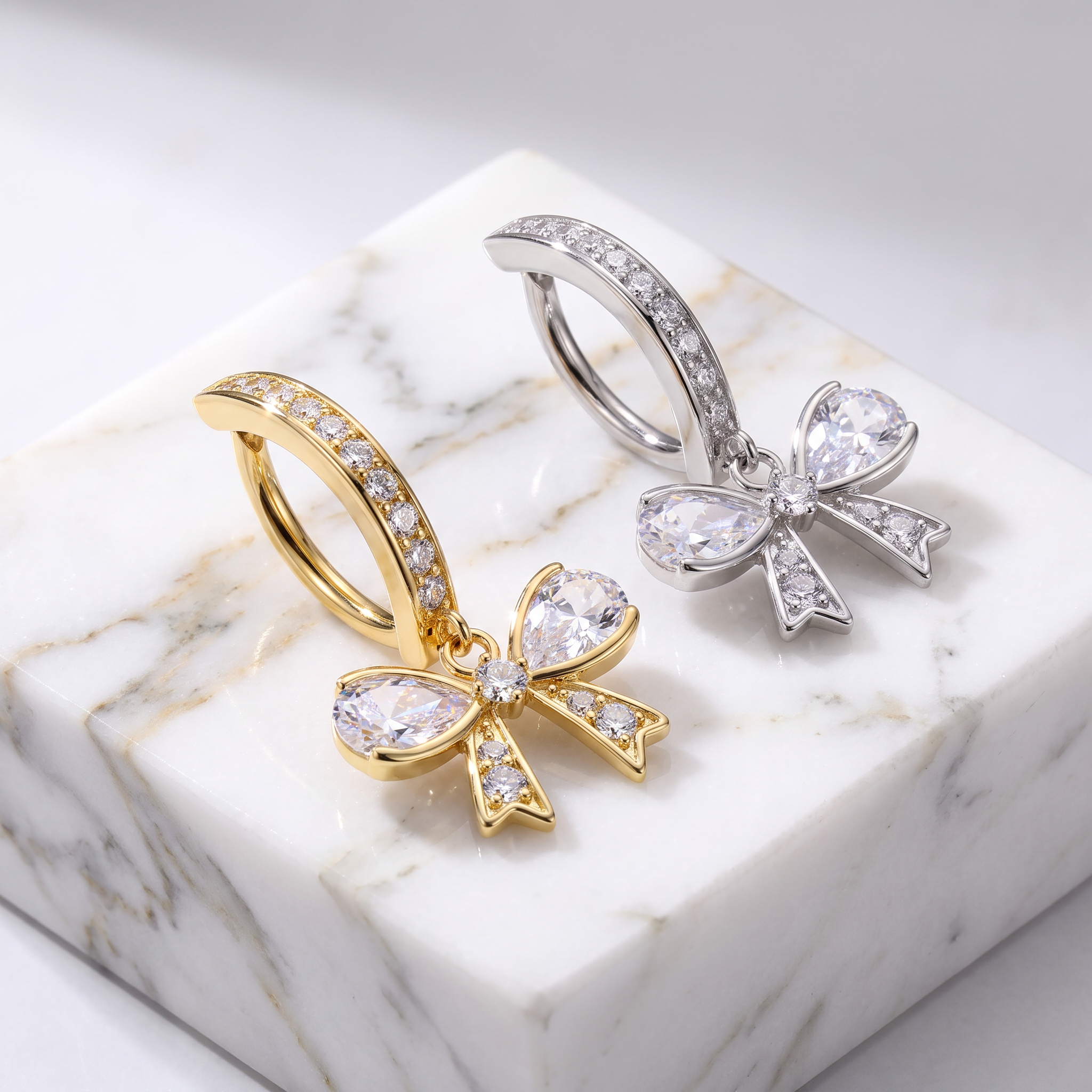 Two bow-shaped hoop earrings displayed on a white marble block. One earring is gold with clear stones, and the other is silver with clear stones.