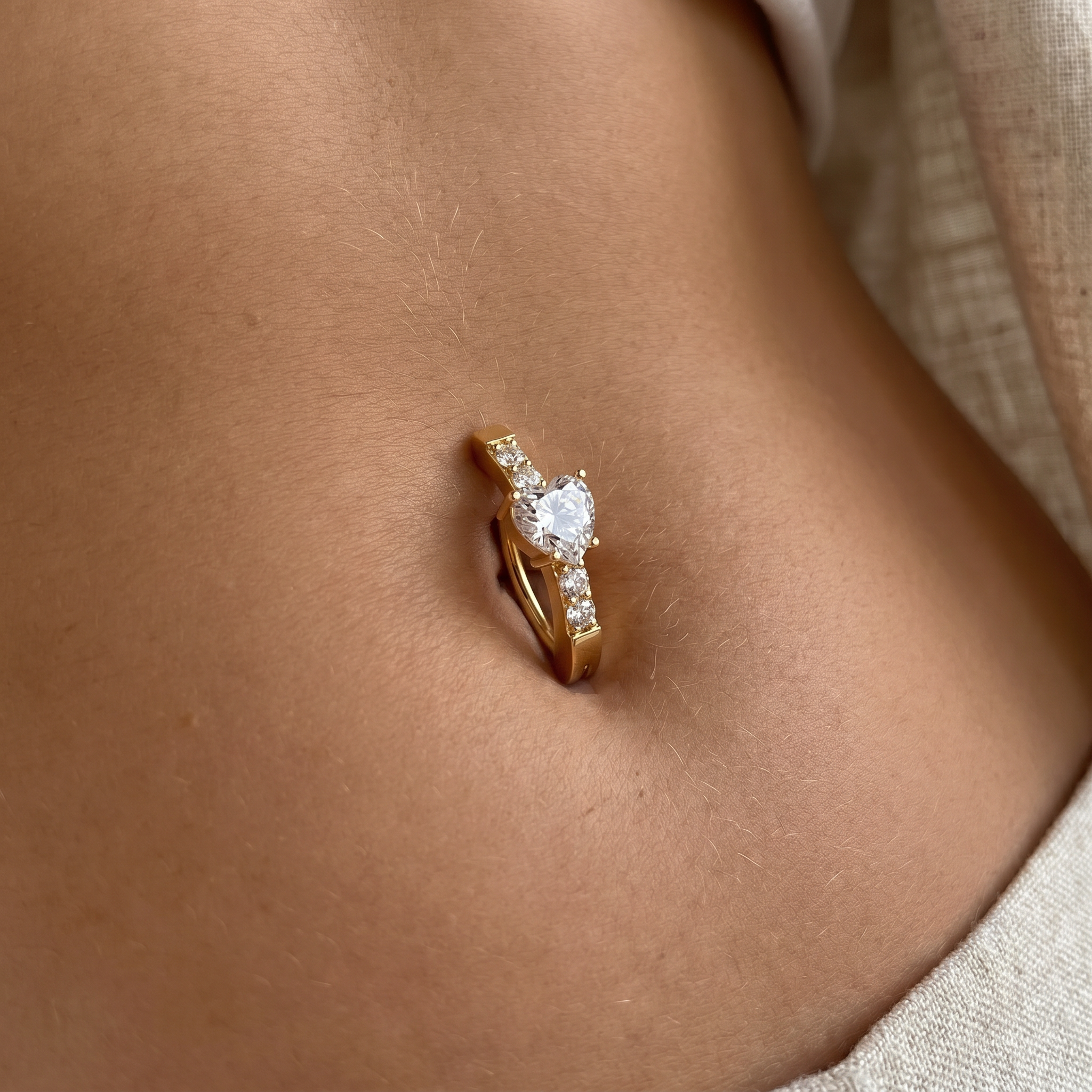 A close-up of a gold belly button ring with a clear heart-shaped gemstone and small side stones, worn on a person's navel.