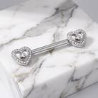 A silver-toned double heart barbell piercing stud with clear pavé-set heart-shaped crystals and a central round crystal, displayed on a white and gray marble block.