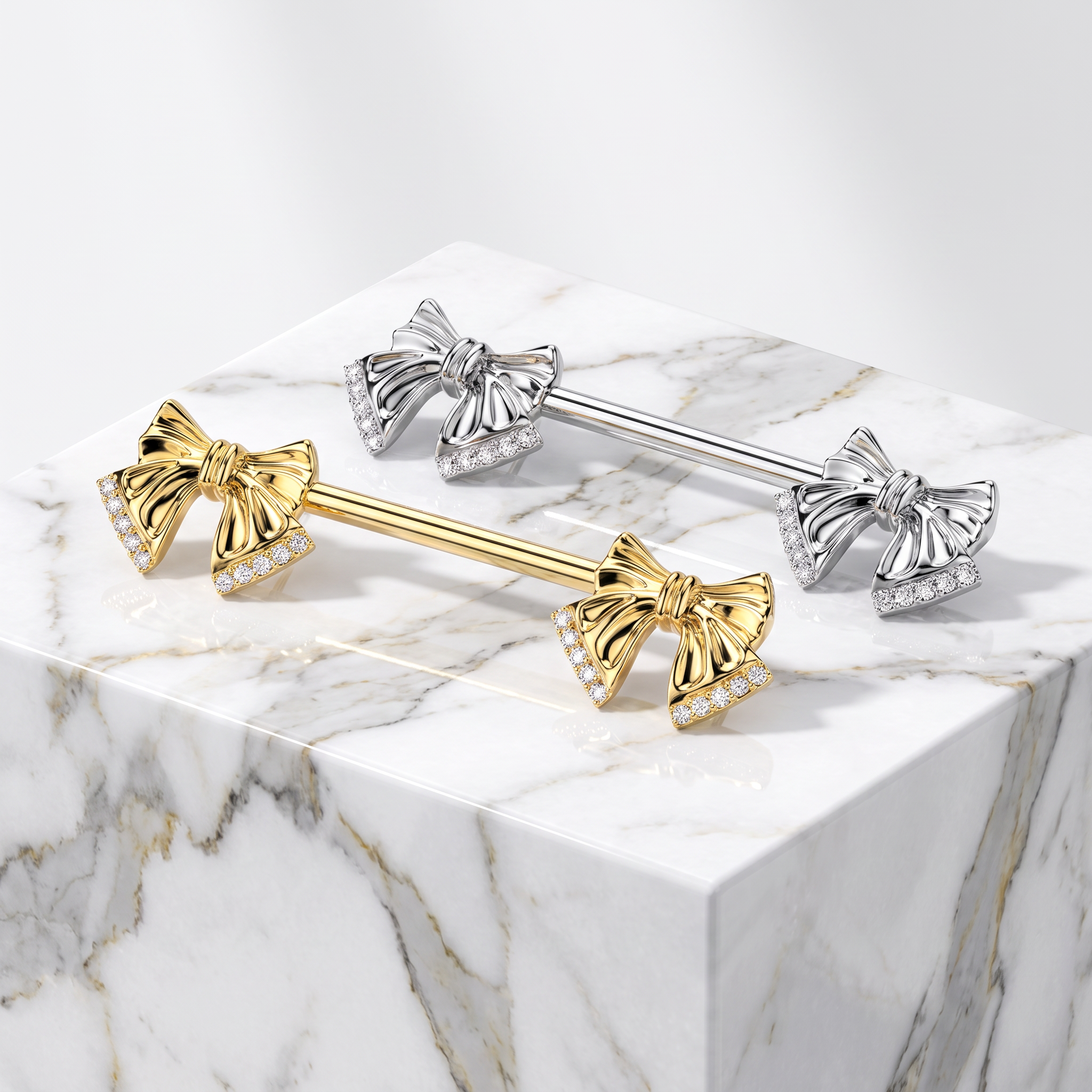 Two barbell-style body jewelry pieces with bow-shaped ends, one in yellow gold and one in silver, both set with small clear stones, displayed on a white marble block against a light gray background.