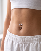 Close-up of a person's midsection wearing a small silver belly button ring with a bow and anchor design, paired with a white ribbed crop top and white shorts.