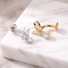 Two butterfly-shaped body jewelry piercings, one in silver and one in gold, both adorned with clear marquise and round crystals, displayed on a white marble block.