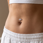 A close-up of a person's lower torso showing a small silver lotus flower belly button ring worn at the navel, paired with a white ribbed crop top and white drawstring shorts.