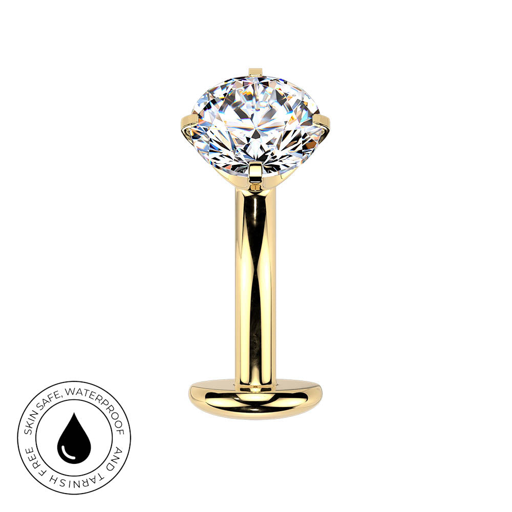 A gold-plated labret piercing stud featuring a large, clear, round-cut gemstone in a four-prong setting. The image is set against a plain white background and includes a circular graphic overlay in the bottom-left corner with a water droplet icon and the text 'SKINSAFE WATERPROOF AND TARNISH FREE'.
