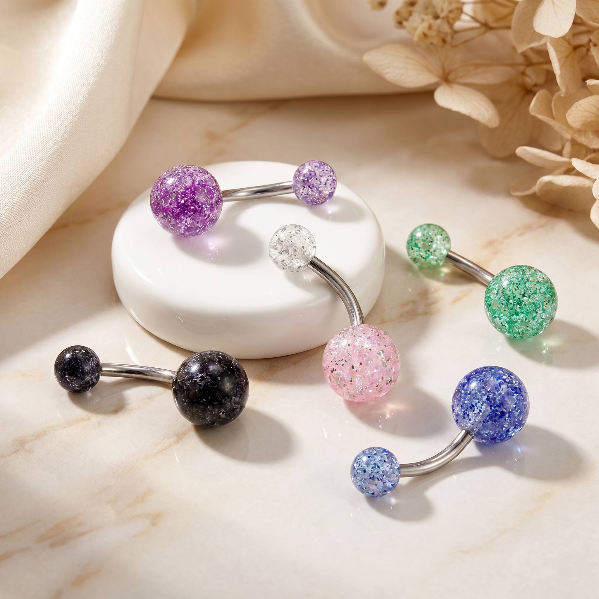 Several curved barbell belly button piercings with glittery gemstone beads are arranged on a light marble surface with a white dish and soft fabric in the background. The visible bead colors include purple, green, pink, blue, black, and clear.
