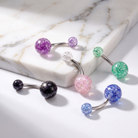 Five curved barbell belly button piercings with glittery spherical beads in purple, clear, green, pink, black, blue, and silver, arranged on a white marble slab and white surface.