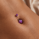A close-up of a person's navel wearing two purple glitter stud and barbell belly button piercings.