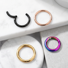 Four metal hoop rings in different finishes displayed on a white marble and gray stone surface: two black rings, one rose gold ring, one gold ring, and one iridescent rainbow ring.