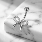 A silver belly button ring with a bow design set with clear crystals, displayed on a white marble block.