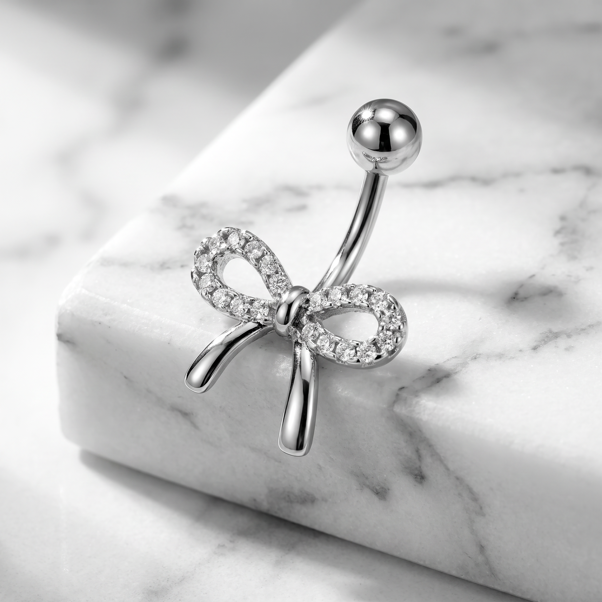 A silver belly button ring with a bow design set with clear crystals, displayed on a white marble block.