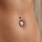 A close-up of a person's lower torso showing a belly button with two silver-toned navel piercings: a small round stud above and a larger heart-shaped halo stud below. The heart stud features a clear teardrop gemstone surrounded by small clear stones.