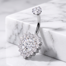 A silver-toned belly button ring with a large marquise-cut clear gemstone surrounded by a halo of smaller clear stones, resting on a white marble block against a white marble surface.