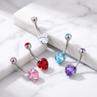 Four silver curved barbell belly button rings with heart-shaped crystals in pink, red, light blue, and purple, displayed on a white marble background.