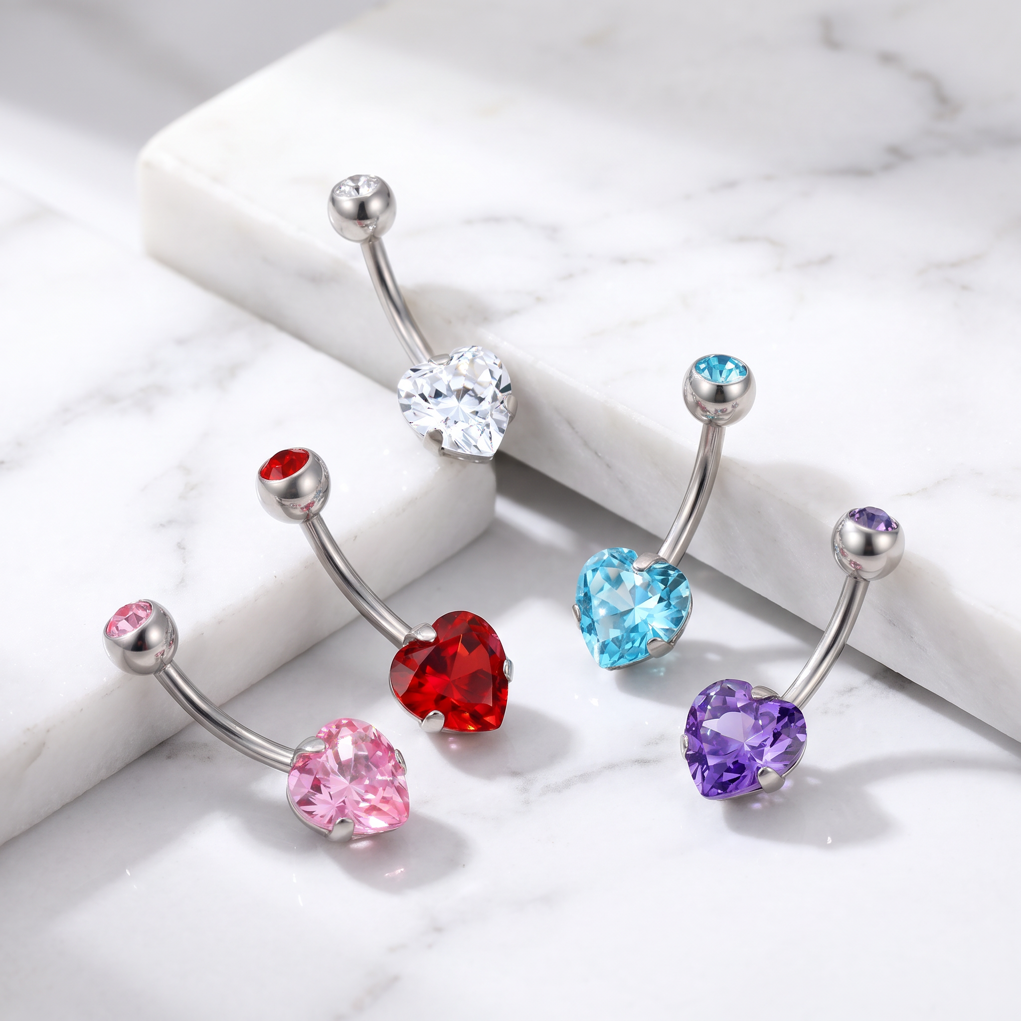Four silver curved barbell belly button rings with heart-shaped crystals in pink, red, light blue, and purple, displayed on a white marble background.