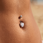 A close-up of a person's lower torso showing a belly button ring with a silver-toned barbell and a clear heart-shaped gemstone, set against warm skin tones and a softly blurred sandy background.