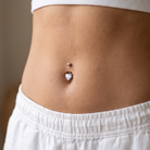 Close-up of a person's lower torso showing a belly button with two piercings: a small silver stud above and a larger silver belly button ring with a clear heart-shaped gemstone below, wearing white shorts.