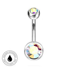 A silver-toned curved barbell belly button ring with a large faceted iridescent crystal on the bottom and a smaller multicolor crystal on the top. A circular badge on the bottom left reads 'SKINSAFE WATERPROOF AND TARNISH FREE'.