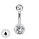 A silver-tone curved barbell navel piercing stud with a large clear round gemstone on the bottom and a smaller clear gemstone on the top, shown on a white background with a circular 'Skin Safe Waterproof' badge in the lower left.