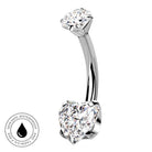 A curved barbell belly button ring with a large heart-shaped clear gemstone on one end and a round clear gemstone on the other, shown on a white background with a small circular skin-safe waterproof badge in the lower left.