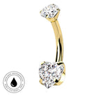 A curved yellow gold navel piercing ring with two clear gemstones, one round and one heart-shaped, shown on a white background with a circular skin-safe waterproof badge in the bottom-left corner.
