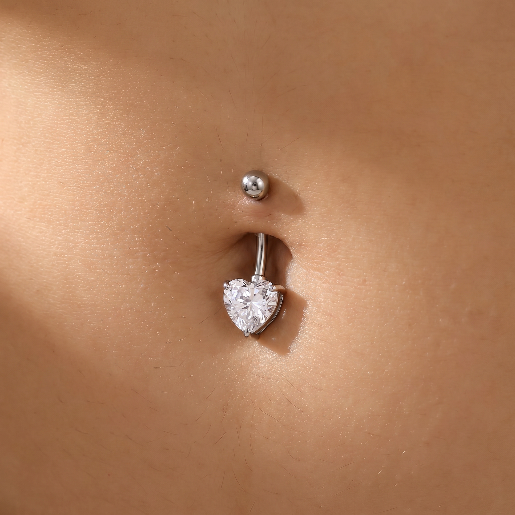 A close-up of a silver belly button ring with a heart-shaped clear gemstone, shown worn on a person's navel.