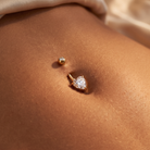 A close-up of a person's navel showing two gold-toned belly button piercings: a small round stud and a larger heart-shaped stud with a clear gemstone.