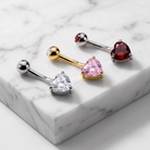 Four heart-shaped barbell piercings displayed on a white marble slab. The set includes silver-toned pieces with clear and white heart stones, a gold-toned piece with a pink heart stone, and a silver-toned piece with a red heart stone.