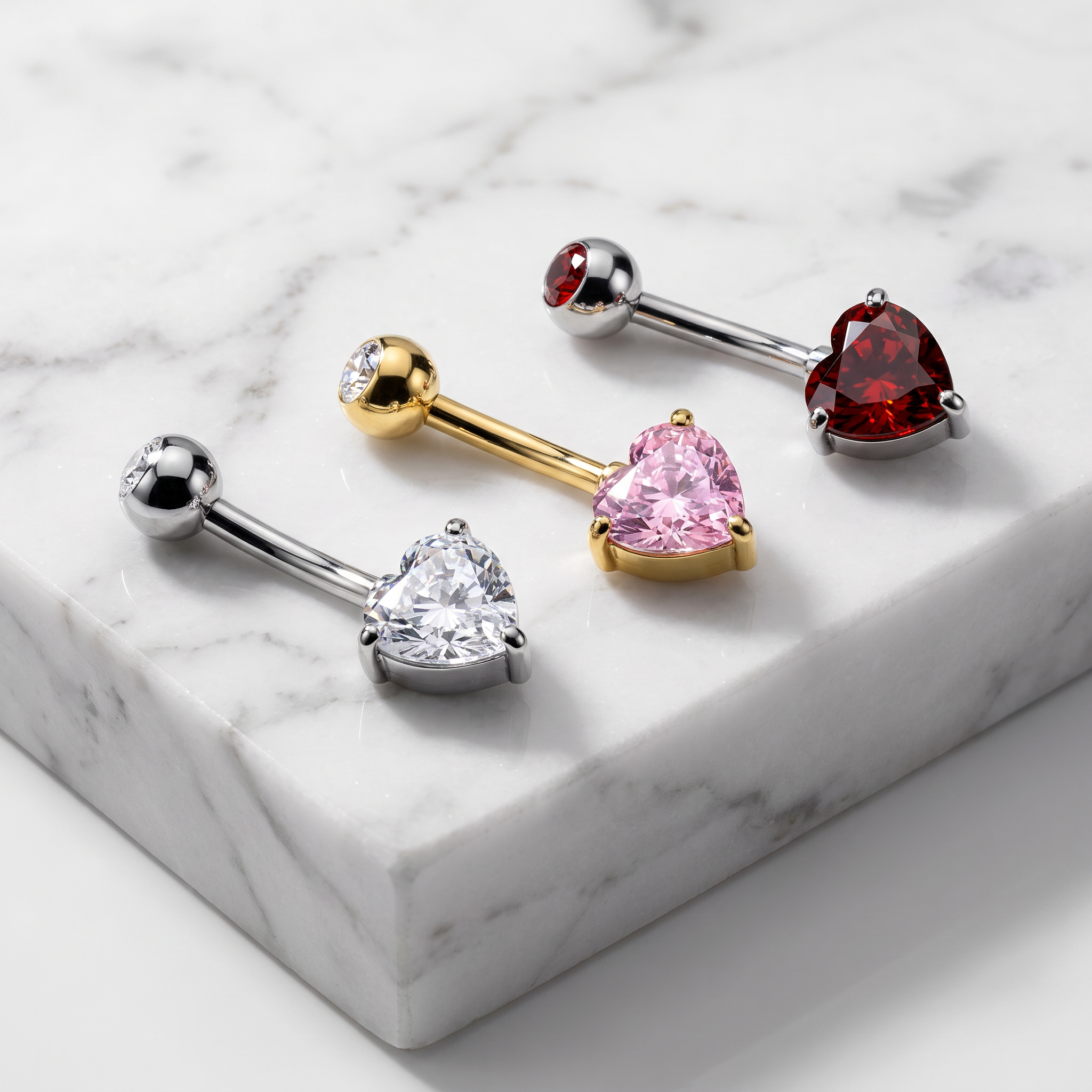 Four heart-shaped barbell piercings displayed on a white marble slab. The set includes silver-toned pieces with clear and white heart stones, a gold-toned piece with a pink heart stone, and a silver-toned piece with a red heart stone.