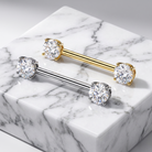 Two barbell-style body jewelry pieces with round clear gemstones at each end, one in yellow gold and one in silver, displayed on a white marble block.