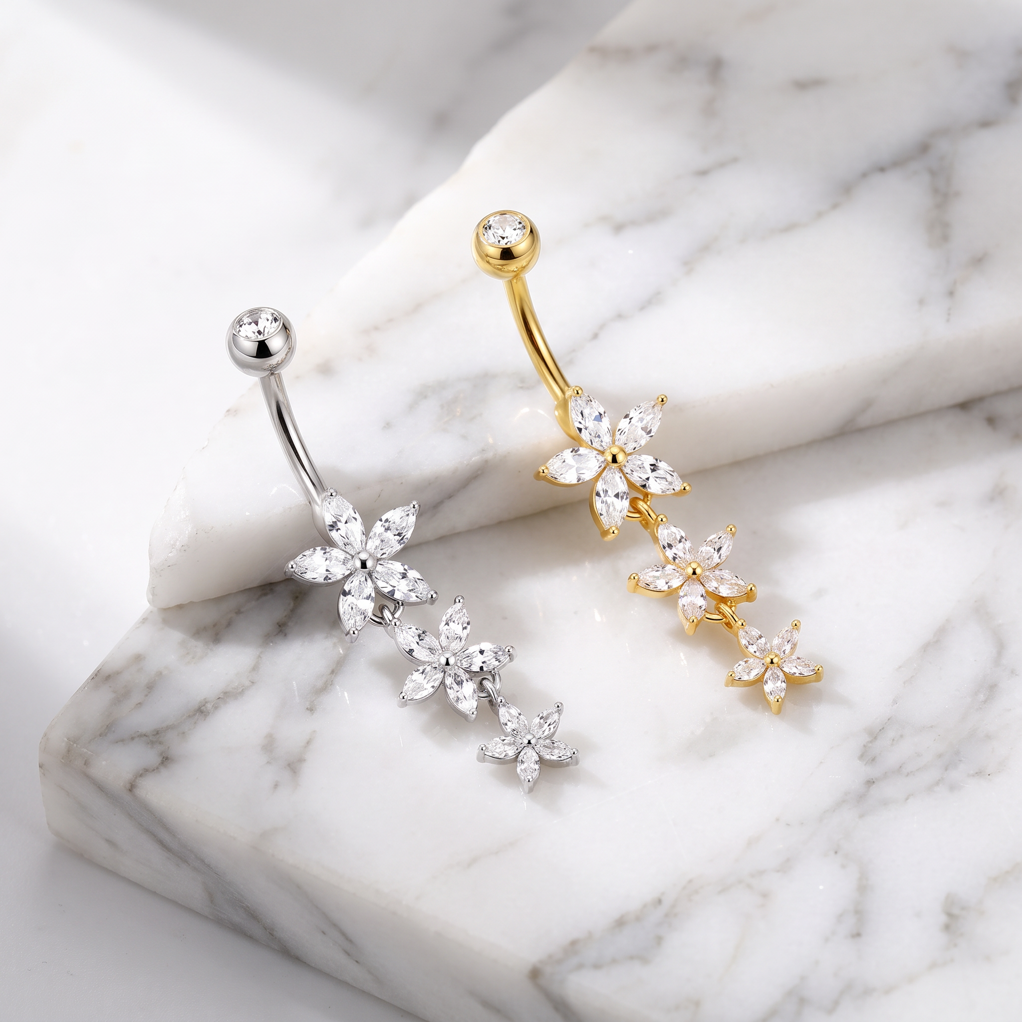 Two flower-shaped belly button rings displayed on a white marble slab. One ring is silver-toned with clear crystal flowers, and the other is gold-toned with clear crystal flowers.