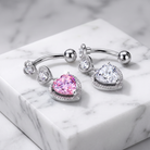 Two silver-toned belly button rings with heart-shaped crystal charms, one pink and one clear, displayed on a white marble block.