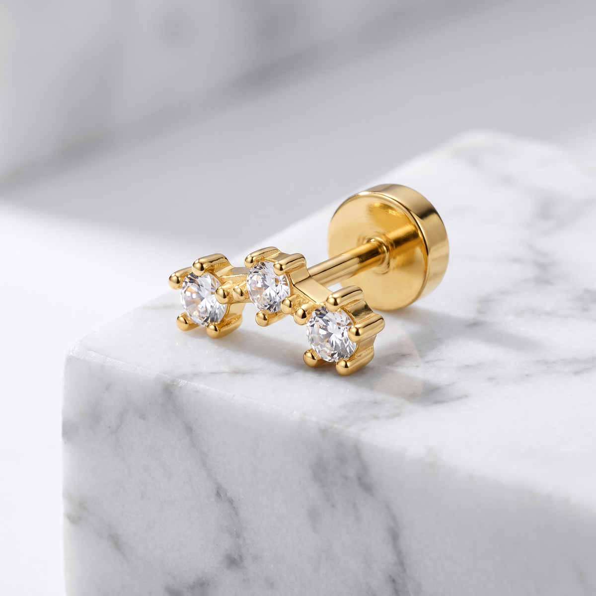 A pair of gold stud earrings with clear gemstones, displayed on a white marble block against a soft gray background.