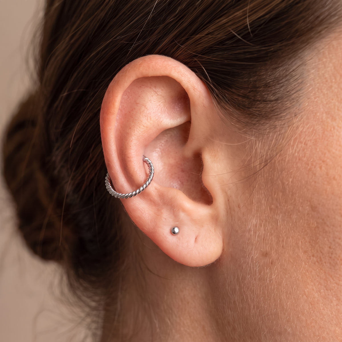 Close-up of a person's ear showcasing two silver ear piercings: a textured hoop cartilage piercing and a small stud lobe piercing.
