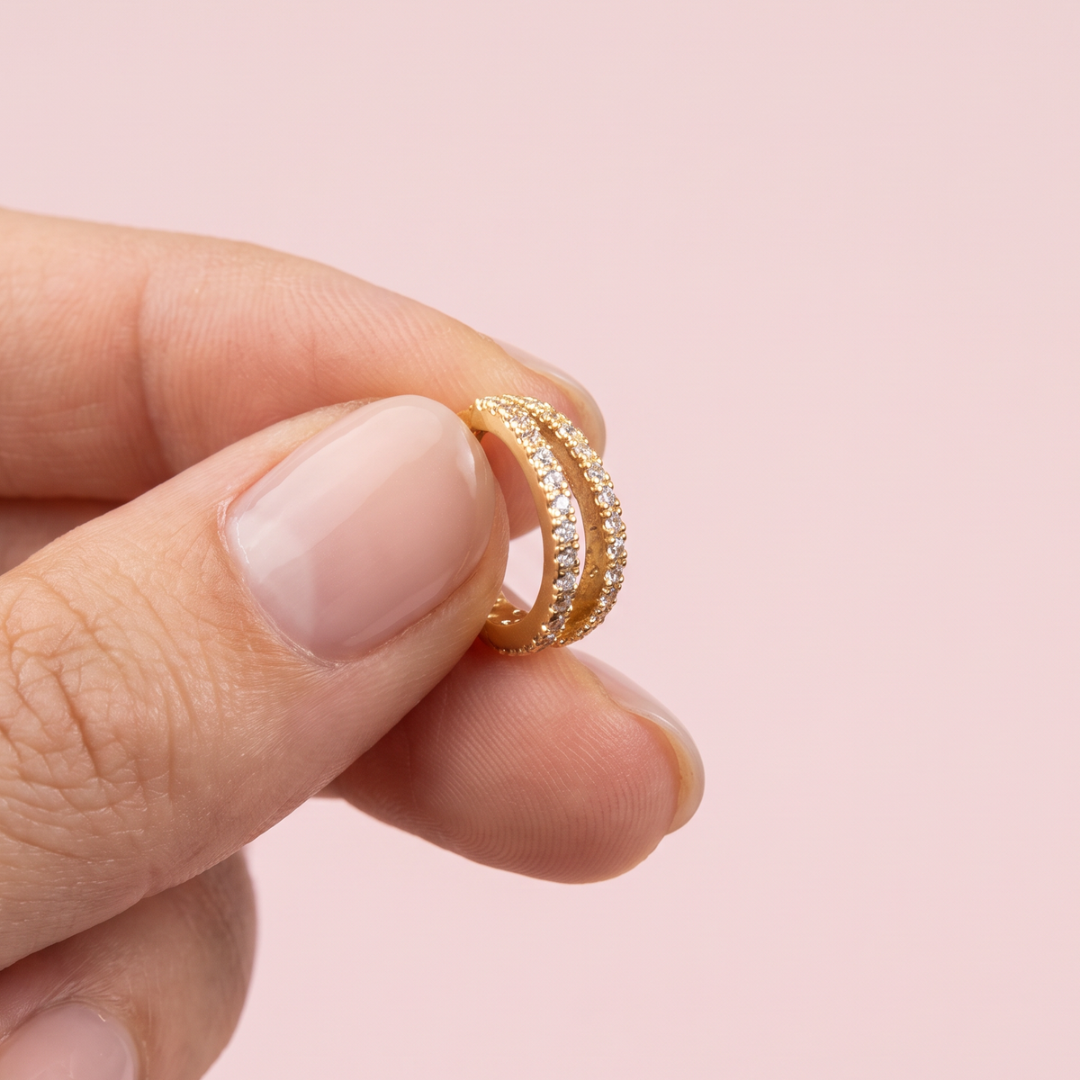 A close-up of a hand holding a delicate gold ring with pavé-set clear stones against a soft pink background.