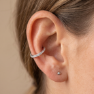 A close-up of a person's ear wearing a silver-toned curved ear cuff adorned with small clear stones, alongside a small stud earring, against a soft beige background.