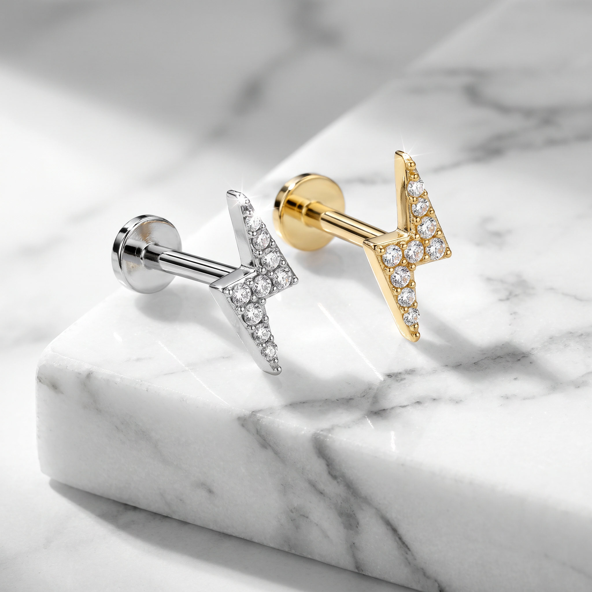 Two lightning bolt stud earrings on a white marble slab, one in silver and one in yellow gold, both set with clear pavé stones.