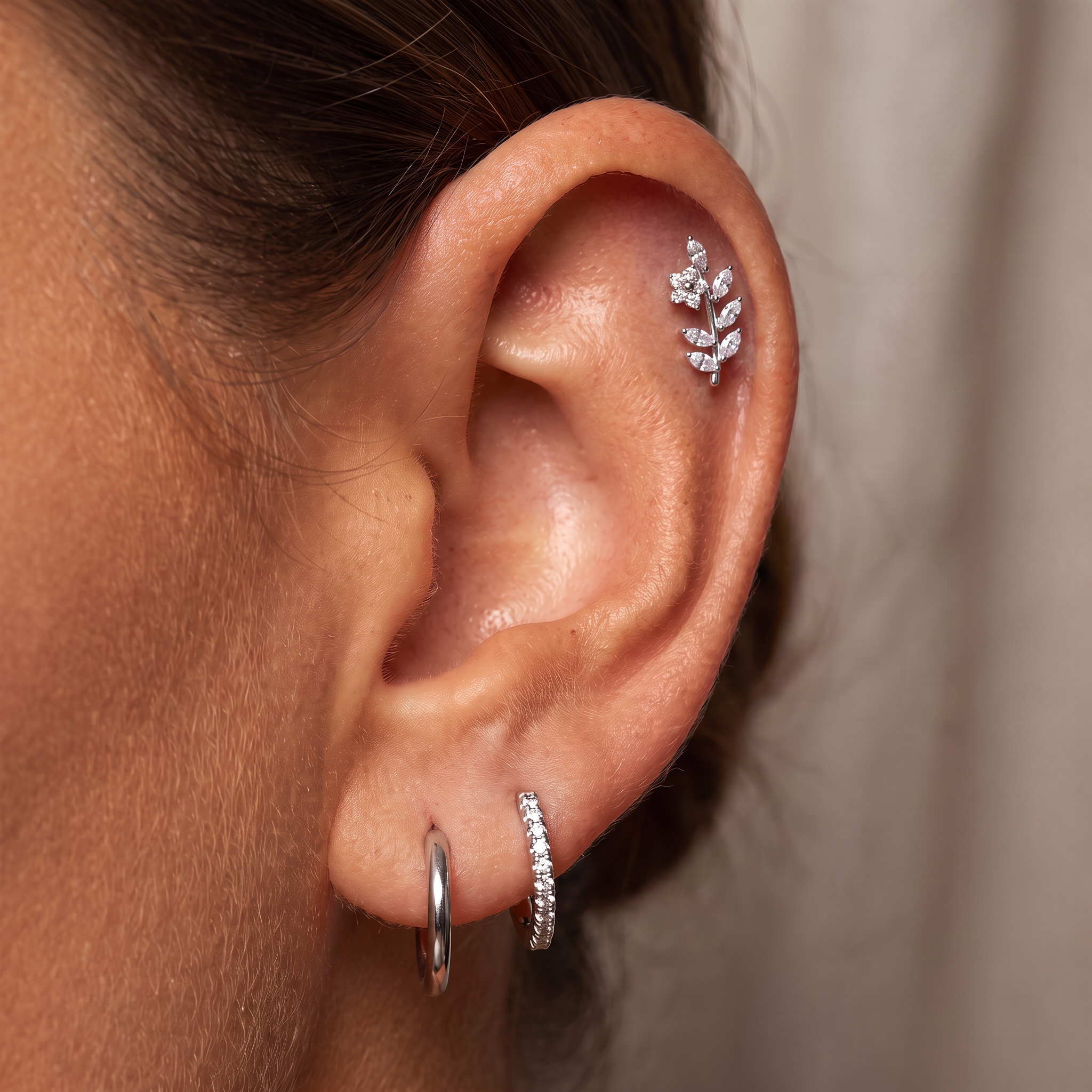 Close-up of a person's ear showcasing three silver ear piercings: a small silver hoop in the lobe, a silver curved hoop set with clear stones on the helix, and a silver leaf-shaped stud set with clear stones on the upper ear cartilage.