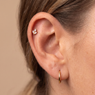 Close-up of a person's ear showcasing two gold ear piercings: a small gold leaf-shaped stud on the upper cartilage and a polished gold hoop in the lobe, against a solid beige background.