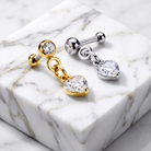 Two heart-shaped labret earrings displayed on a white marble block. One earring is gold-toned with a heart-shaped clear gemstone, and the other is silver-toned with a matching heart-shaped clear gemstone.