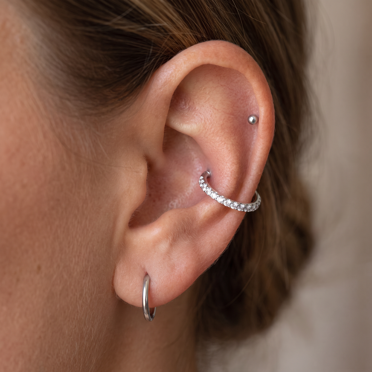 Close-up of a person's ear showcasing silver body jewelry, including a silver hoop in the lobe, a silver cartilage hoop adorned with small clear stones, and a silver dome stud in the rib cage.