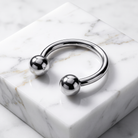 A polished silver-tone metal cuff bracelet with two spherical ball ends, displayed on a white marble block over a marble surface.