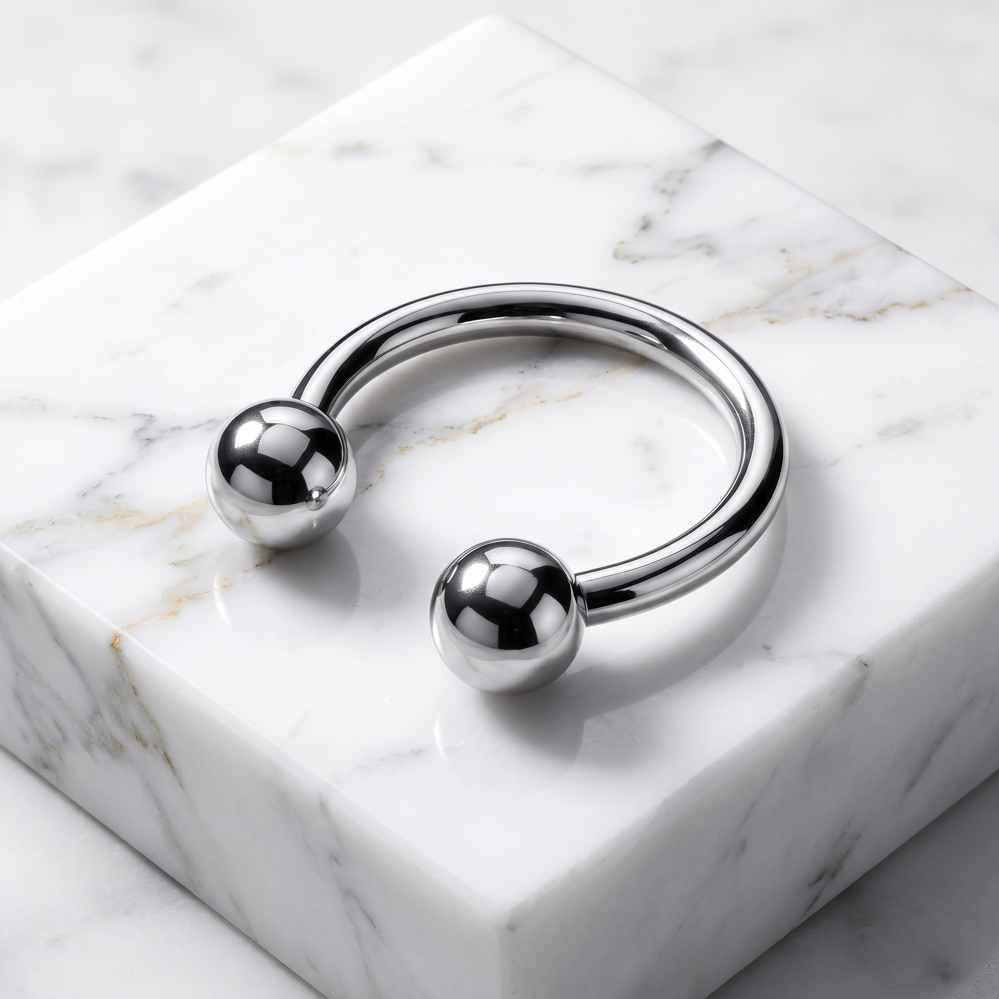 A polished silver-tone metal cuff bracelet with two spherical ball ends, displayed on a white marble block over a marble surface.