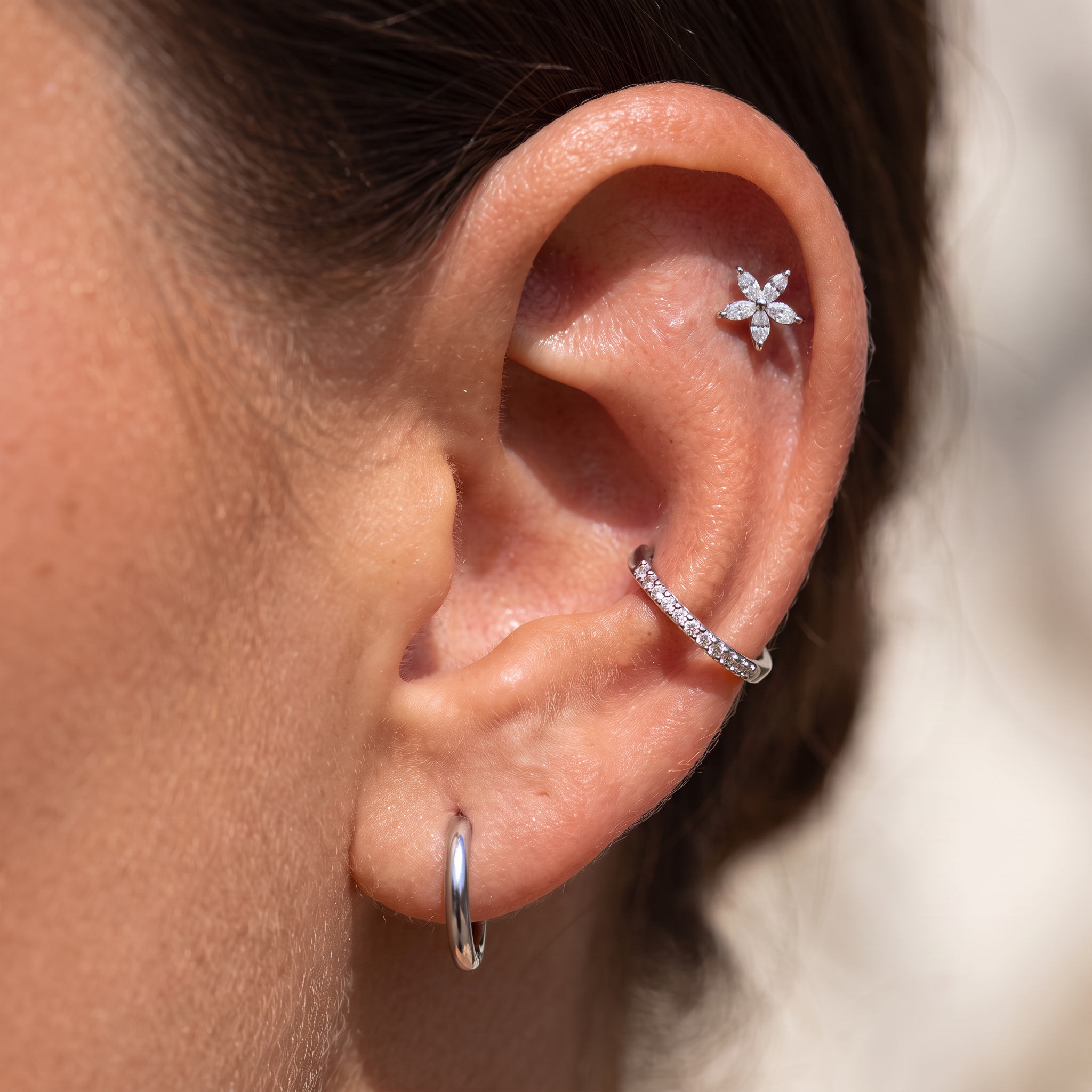 Close-up of a person's ear showcasing multiple silver-toned ear piercings: a small flower-shaped stud in the lobe, a curved huggie hoop along the upper ear cartilage, and a simple hoop in the lobe.